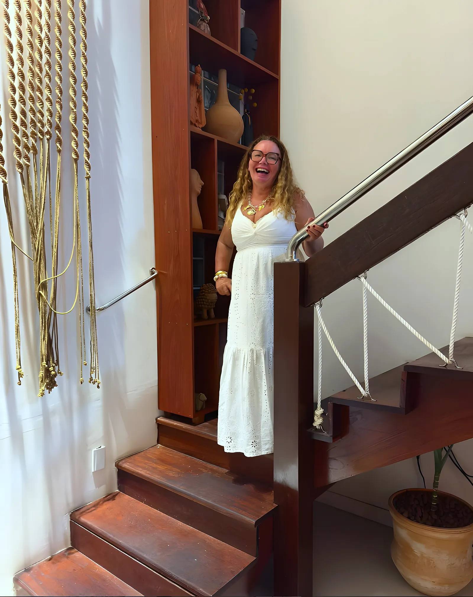 Nilda Carneiro, fundadora da Nilda Cortinas, sorrindo de vestido branco na escada de madeira, em Feira de Santana